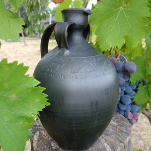 Può includere: Una brocca in ceramica nera con disegni intricati e due manici, appoggiata su una superficie di legno con un grappolo d'uva sullo sfondo.