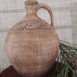 May include: A brown ceramic pitcher with a carved design and a handle. The pitcher is sitting on a woven surface with a pine branch and a pine cone.