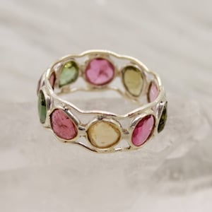 Puede incluir: Un anillo de plata con una banda de pequeñas piedras preciosas redondas multicolores. Las piedras preciosas están engastadas en un patrón ondulado y son rosas, verdes y amarillas.