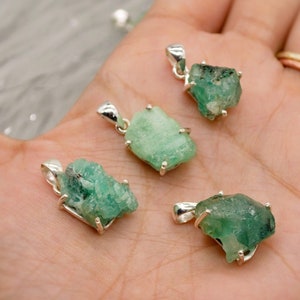 May include: Four raw emerald gemstone pendants with silver settings. The emeralds are a deep green color and have a rough, natural texture. The pendants are all different sizes and shapes.