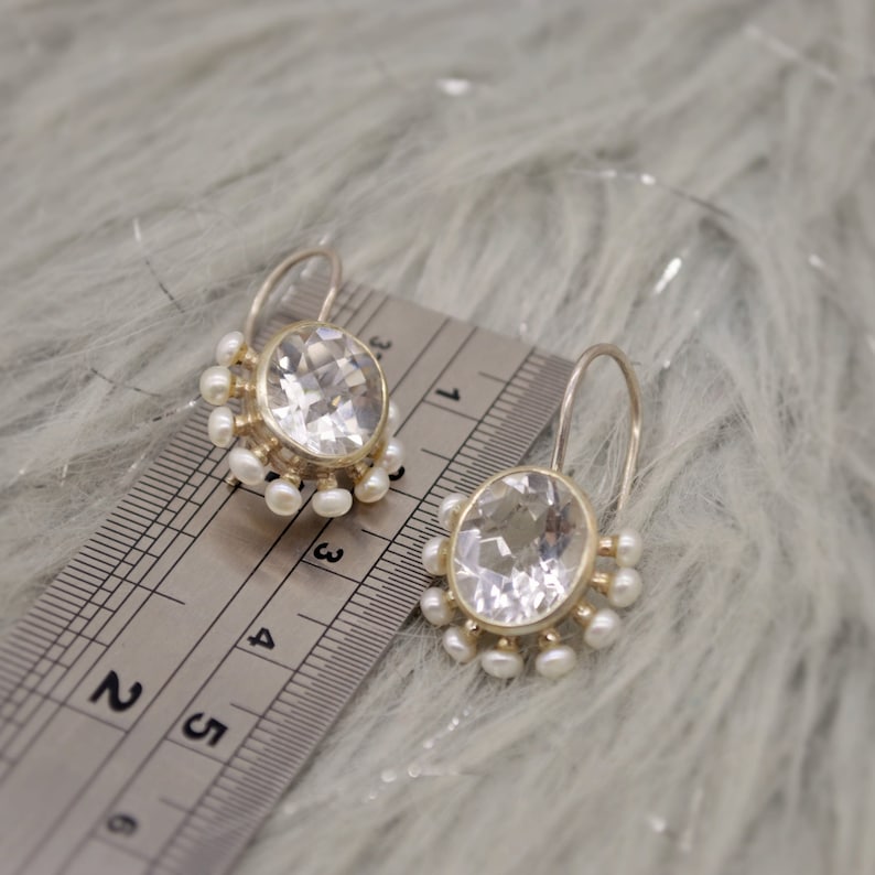 May include: A pair of silver-toned earrings featuring a large, oval, faceted clear stone surrounded by small white pearls. A metal ruler is placed next to the earrings, showing the stones measure approximately 0.5 inches in width.