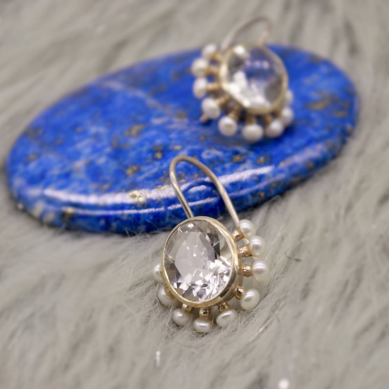 May include: A pair of drop earrings featuring a large, round, faceted clear stone set in a gold-toned metal frame. The perimeter of each setting is adorned with a series of small, white, spherical pearls. Each earring includes a curved wire hook.