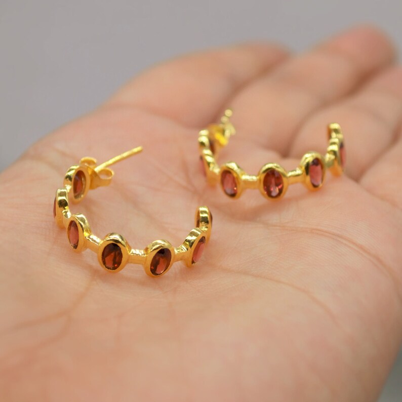 May include: Gold hoop earrings with red gemstones. The earrings are a half circle shape and have a total of six gemstones.