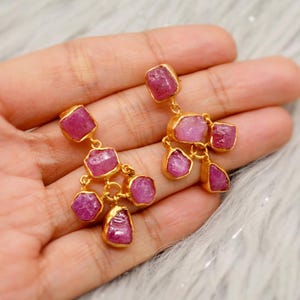 May include: A pair of gold earrings with pink ruby gemstones. The earrings are dangle style and feature multiple ruby stones of varying sizes.