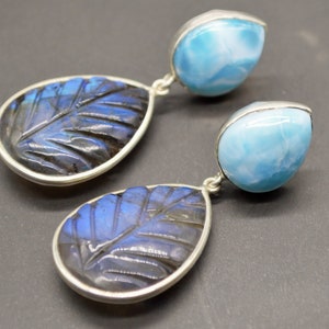 May include: A pair of dangle earrings featuring a carved labradorite stone and a blue sky agate stone. The earrings are silver toned.