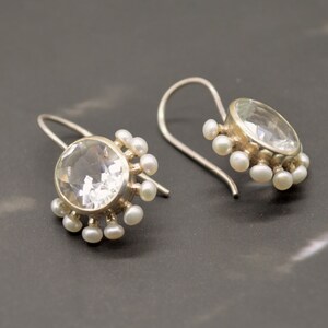 May include: A pair of silver drop earrings featuring a large, round, faceted clear gemstone in the center. The setting is surrounded by a halo of small, white, round pearls. Each earring is attached to a curved silver ear wire for hanging.