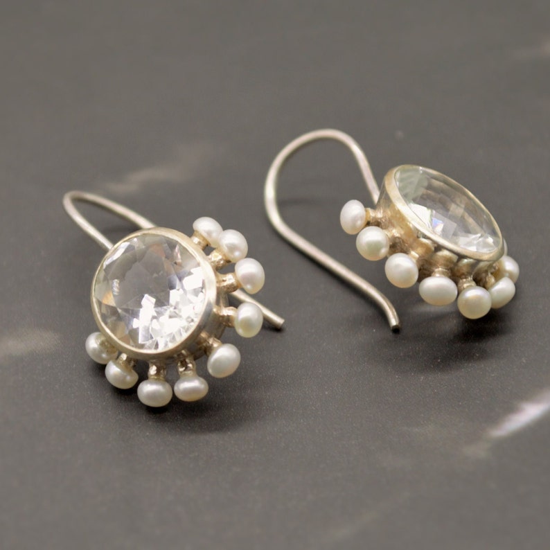 May include: A pair of silver drop earrings featuring a large, round, faceted clear gemstone in the center. The setting is surrounded by a halo of small, white, round pearls. Each earring is attached to a curved silver ear wire for hanging.