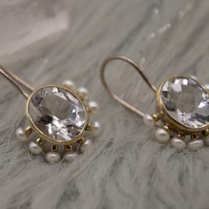 May include: A pair of gold-toned drop earrings featuring a large, oval-cut, clear faceted gemstone at the center. The gemstone is surrounded by a halo of small, round, white pearls attached to the base of the setting, with curved ear wires.