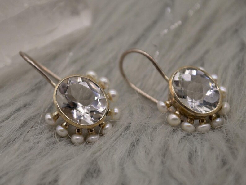May include: A pair of gold-toned drop earrings featuring a large, oval-cut, clear faceted gemstone at the center. The gemstone is surrounded by a halo of small, round, white pearls attached to the base of the setting, with curved ear wires.