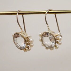 May include: A pair of silver drop earrings featuring a large, round, faceted clear stone in the center. The perimeter of each setting is adorned with a series of small, round, white pearls. The earrings hang from simple, curved silver ear wires.