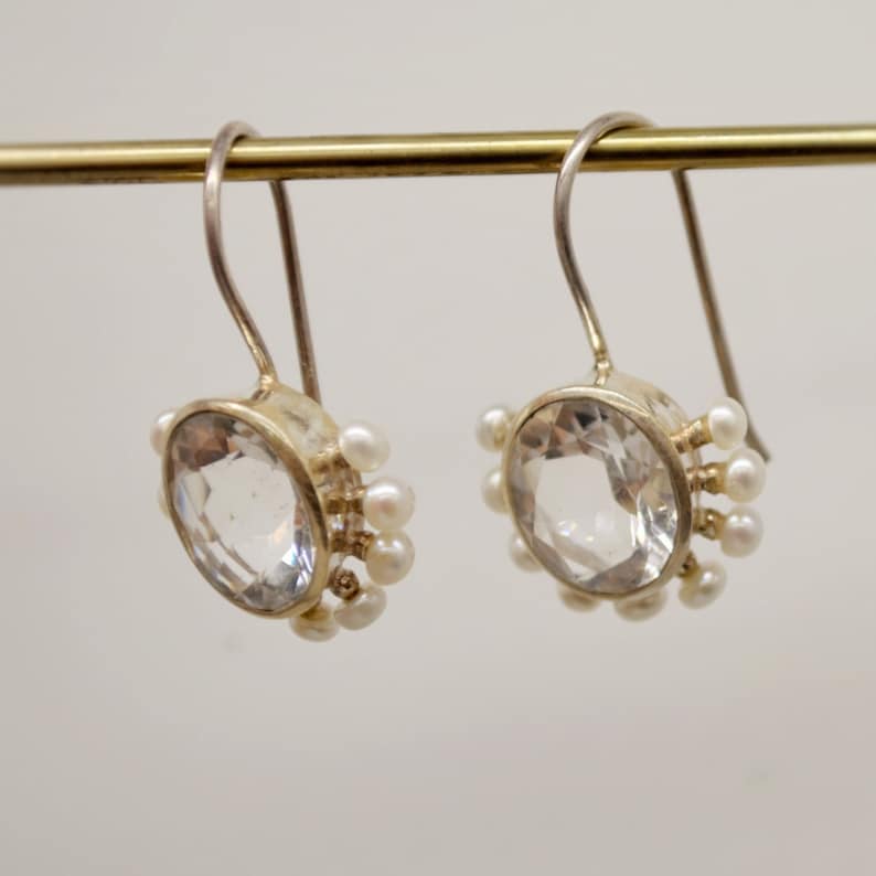 May include: A pair of silver drop earrings featuring a large, round, faceted clear stone in the center. The perimeter of each setting is adorned with a series of small, round, white pearls. The earrings hang from simple, curved silver ear wires.