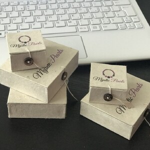 May include: Four square, off-white textured paper gift boxes stacked in two groups. Each box features a dark brown button closure with a string loop and the printed logo Mystic Pearls, which includes a graphic of a beaded necklace.