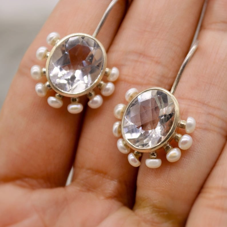 May include: A pair of drop earrings featuring oval-shaped, faceted clear gemstones set in silver-toned metal. Each central stone is surrounded by a series of small, round white pearls arranged along the lower edge, attached to silver ear wires.