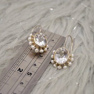 May include: A pair of silver earrings featuring oval faceted clear stones surrounded by small white pearls. The earrings are positioned next to a metal ruler showing a measurement of approximately 0.75 inches for the stone setting width.