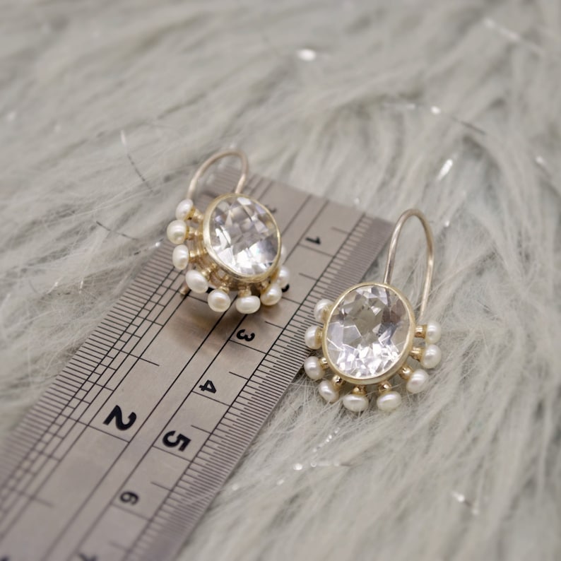 May include: A pair of silver earrings featuring oval faceted clear stones surrounded by small white pearls. The earrings are positioned next to a metal ruler showing a measurement of approximately 0.75 inches for the stone setting width.