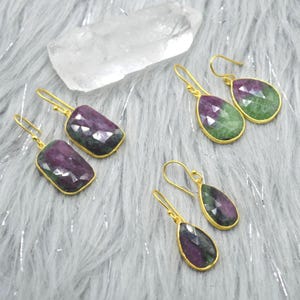 May include: Gold-toned earrings with faceted, multi-colored gemstones. The earrings feature a rectangular and a teardrop shape, with a gold frame. The gemstones are a mix of purple and green hues. The earrings are displayed on a soft, gray surface.