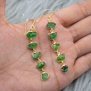 May include: Gold-tone earrings featuring a series of five raw, green gemstones. The gemstones are irregularly shaped and set in gold-colored bezels. The earrings have a hook closure.
