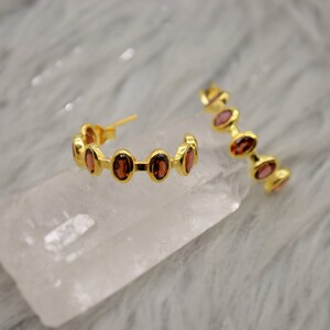 May include: A pair of gold hoop earrings with red gemstones. The earrings are on a white surface.