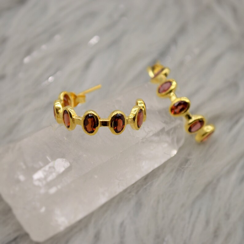 May include: A pair of gold hoop earrings with red gemstones. The earrings are on a white surface.