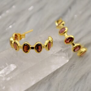 May include: Gold hoop earrings with a row of oval-shaped red gemstones.