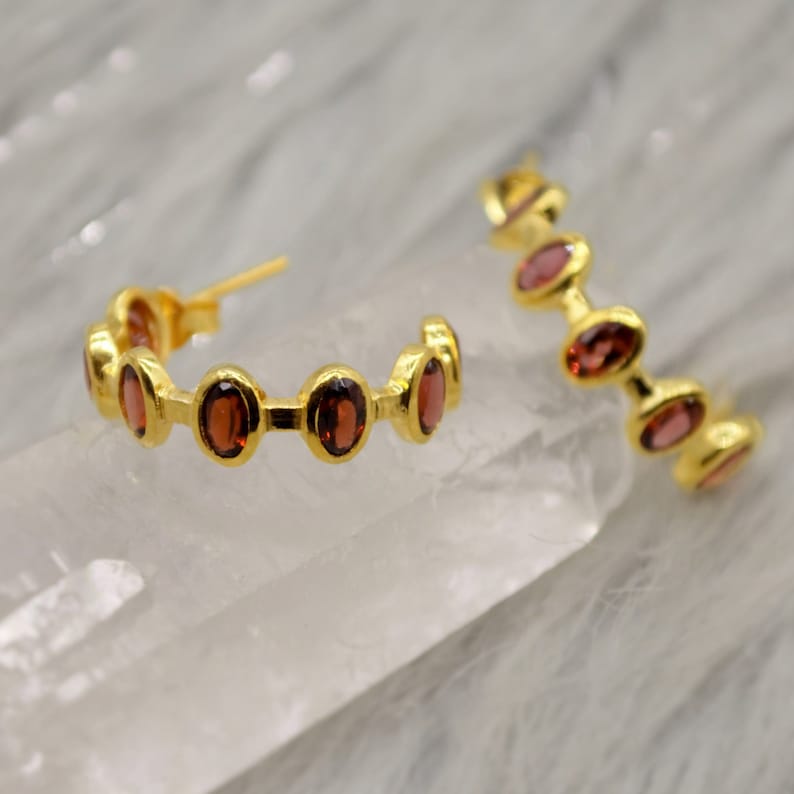 May include: Gold hoop earrings with a row of oval-shaped red gemstones.
