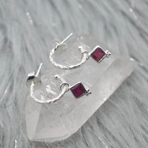 Peut inclure: Paire de boucles d'oreilles créoles en argent avec une finition texturée. Chaque boucle d'oreille est ornée d'un pendentif carré en pierre précieuse rouge foncé. Présentées sur une surface cristalline transparente.