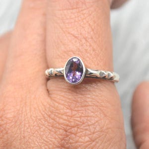 Könnte beinhalten: Ein silberner Ring mit einem ovalen, tiefvioletten Edelstein. Das Band hat ein strukturiertes Muster. Der Ring wird an einem Finger gezeigt.