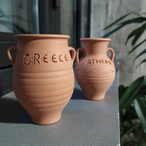 May include: Two terracotta pots with the words "GREECE" and "ATHENS" inscribed on them. The pots are sitting on a gray surface.