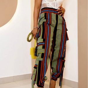 May include: A white lace bodysuit paired with high-waisted cargo pants featuring vertical stripes in red, blue, yellow, and black. The pants have multiple pockets and tie details. The outfit is completed with gold heels and a yellow beaded handbag.