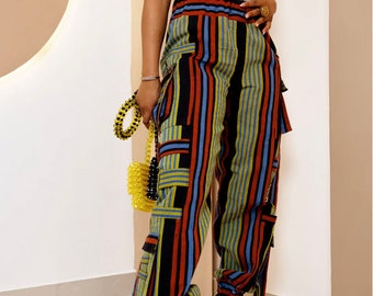 May include: A white lace bodysuit paired with high-waisted cargo pants featuring vertical stripes in red, blue, yellow, and black. The pants have multiple pockets and tie details. The outfit is completed with gold heels and a yellow beaded handbag.