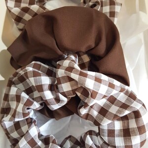 May include: Three brown scrunchies in a white box. Two scrunchies feature a brown and white gingham pattern, and one is plain brown. The scrunchies are made of soft fabric, suitable for various hairstyles.