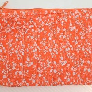 May include: An orange quilted clutch bag with a white floral design. The rectangular bag features a zip closure and a small wrist strap. The fabric is patterned with white flowers and vines. The zip pull is orange.