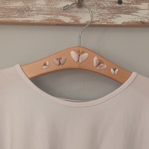 May include: A wooden clothes hanger with a white shirt hanging on it. The hanger is decorated with several small pink seashells that are arranged in a butterfly shape.