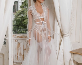 Può includere: Un completo intimo bianco trasparente, composto da vestaglia, reggiseno e slip a vita alta. La vestaglia presenta maniche lunghe e delicati dettagli in pizzo sulle spalle e sui polsini. Intimo davanti a una finestra con tende bianche.