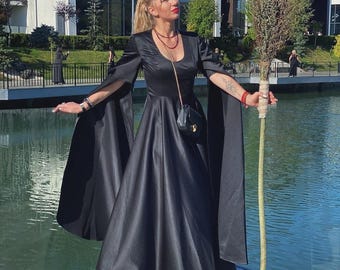 May include: A black satin gown with long, flowing sleeves and a full skirt. The dress features a scoop neckline and is accessorized with a small black purse. The woman is holding a wooden staff. The background includes a body of water and a modern building.
