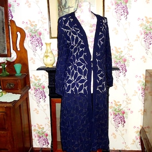 Puede incluir: Un cárdigan largo de encaje azul marino con un diseño de hojas se muestra en un maniquí. El diseño abierto revela una capa inferior blanca. El cárdigan llega hasta el suelo, y la imagen está ambientada en una habitación con papel pintado floral y muebles antiguos.