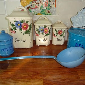 Op de afbeelding: Een verzameling vintage keukengerei, waaronder crèmekleurige voorraadpotten met bloemmotieven en de labels "Sucre", "Café" en "Thé". Een blauwe kruidenpot met het label "POIVRE", een blauwe zoutcontainer met het label "SEL" en een blauwe pollepel.