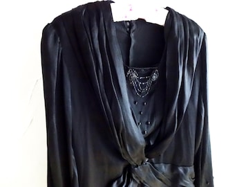 Op de afbeelding: Een zwarte vintage jurk met lange mouwen en een gedrapeerde halslijn. De jurk heeft een kralenversiering op het lijfje en een geplooid taille detail. De jurk hangt aan een witte hanger tegen een witte achtergrond.