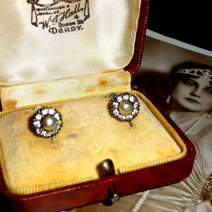 May include: Vintage pearl and rhinestone earrings displayed in a red jewelry box with a yellow velvet interior. The earrings have a central pearl surrounded by a halo of sparkling rhinestones. A vintage photograph is in the background.