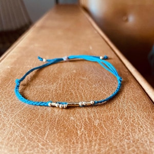 Puede incluir: Un brazalete de cuerda trenzada azul con cuentas plateadas y un dije de barra plateada.