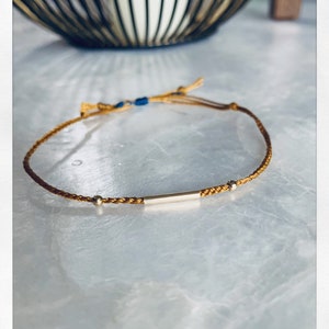 May include: A gold bracelet with a thin gold bar and two small gold beads. The bracelet is made of a braided brown cord.