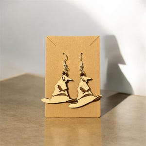 May include: A pair of wooden earrings shaped like the Sorting Hat from the Harry Potter series. The earrings are brown and have a silver hook.