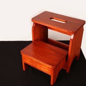 May include: A two-step wooden step stool with a rich, reddish-brown finish. The top step has a handle cutout. The stool is designed with a smaller step below. The background is white and black.