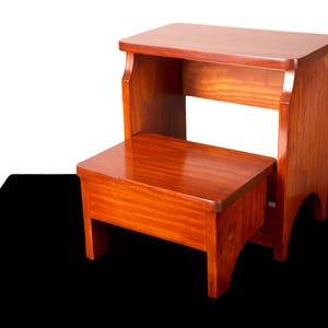 Puede incluir: Un taburete de madera de dos escalones con un rico acabado marrón rojizo. El taburete tiene una parte superior rectangular y un escalón inferior, ambos con bordes redondeados. El diseño es simple y funcional.