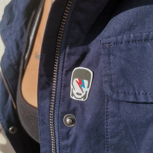 May include: A navy blue jacket with a silver zipper and a silver button. A rectangular pin featuring a stylized image of a person with a lightning bolt on the face is attached to the jacket.
