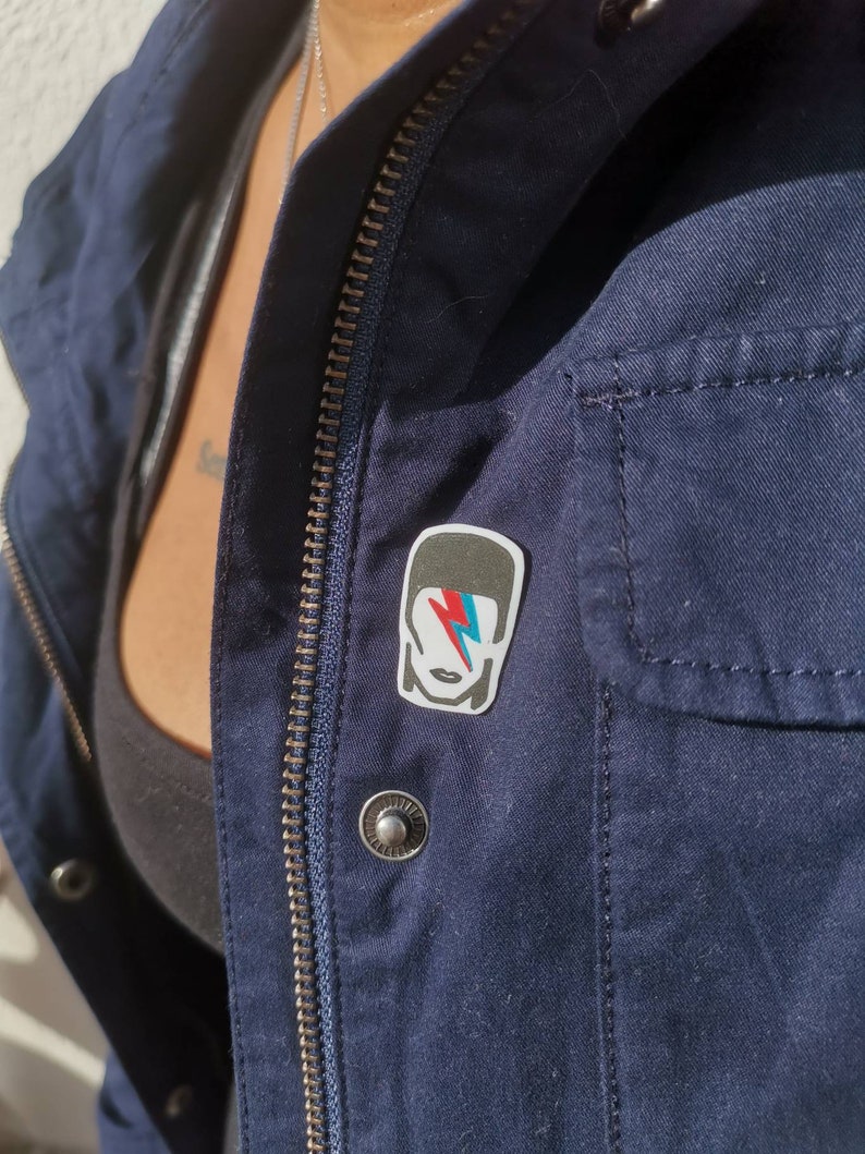 May include: A navy blue jacket with a silver zipper and a silver button. A rectangular pin featuring a stylized image of a person with a lightning bolt on the face is attached to the jacket.