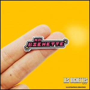 Peut inclure: Une broche émaillée rectangulaire portant le texte MA BICHETTE en lettres capitales roses avec un contour noir. Le design comprend deux petites étoiles jaunes sur les côtés gauche et droit, sur un fond blanc avec une fine bordure noire.