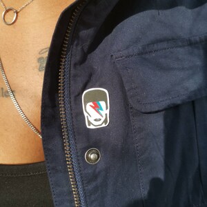 May include: A navy blue jacket with a silver zipper and a small pin featuring a stylized image of a person with a lightning bolt on their face. The pin is attached to the jacket near the zipper.