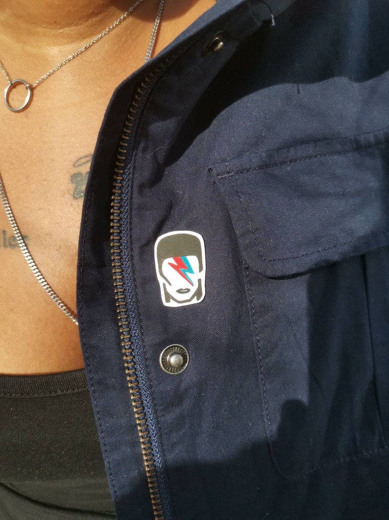 May include: A navy blue jacket with a silver zipper and a small pin featuring a stylized image of a person with a lightning bolt on their face. The pin is attached to the jacket near the zipper.