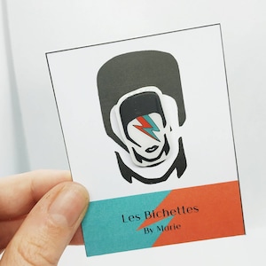 May include: A white card featuring a black and white illustration of a person with a lightning bolt across the face. The lightning bolt is red and blue. The card has the text "Les Bichettes By Marie" in the bottom corner.
